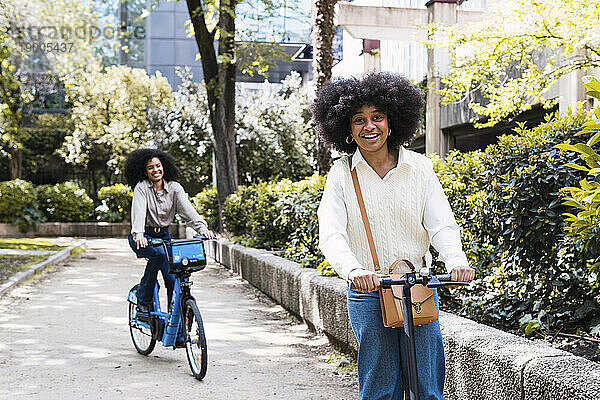 Fröhliche junge Freunde  die gemeinsam im Park Elektroroller und Fahrrad fahren