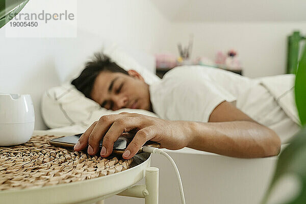 Tired man keeping smart phone on side table near bed