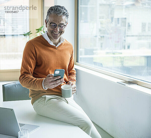 Lächelnder Geschäftsmann mit Smartphone am Tisch im Büro sitzend