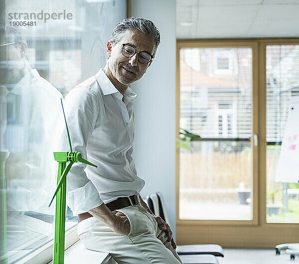 Lächelnder reifer Geschäftsmann blickt auf das Windturbinenmodell  das auf der Fensterbank im Büro lehnt