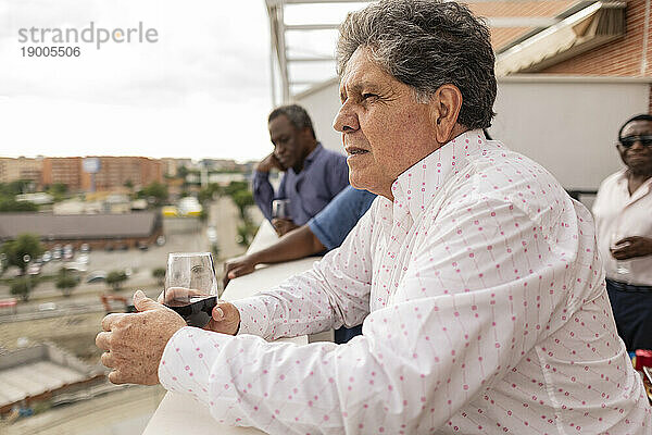 Nachdenklicher älterer Mann lehnt an der Wand und hält ein Weinglas auf dem Balkon