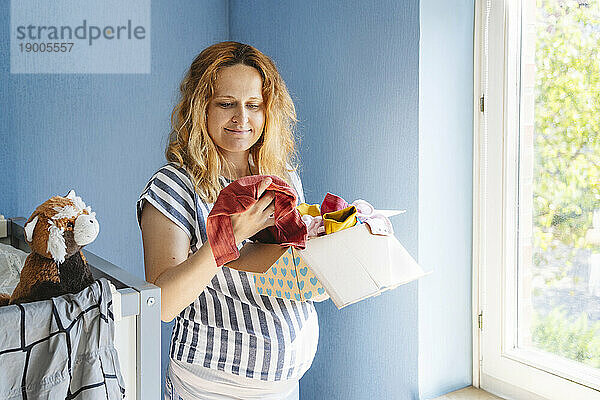 Lächelnde schwangere Frau hält eine Schachtel mit Babykleidung in der Hand