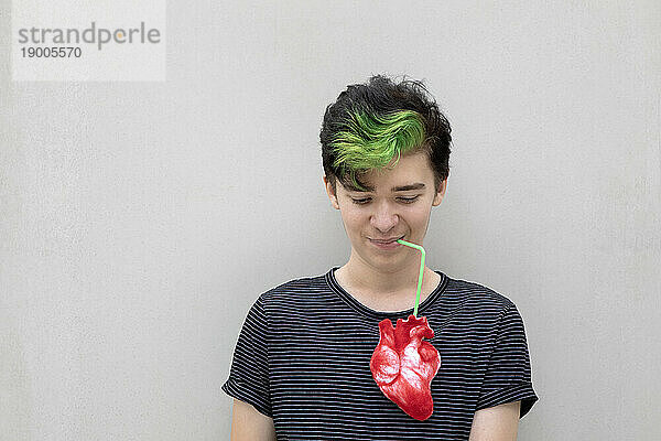 Teenage boy with dyed green hair drinking from heart against gray background