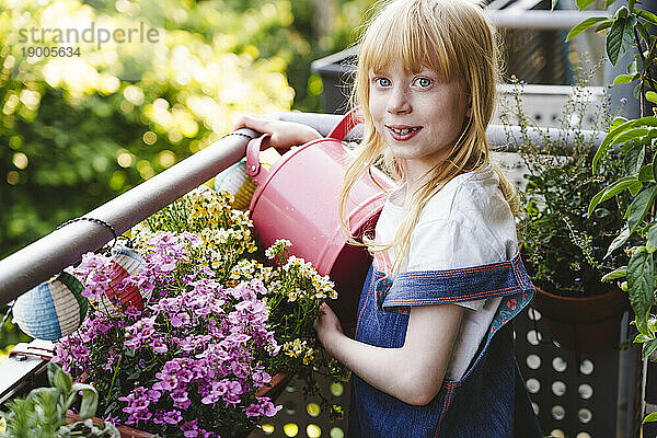 Lächelndes Mädchen mit Gießkanne steht neben Blumentopf auf dem Balkon