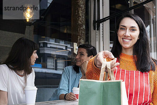 Lächelnde Frau hält eine Papiertüte in der Hand  während sich Freunde im Hintergrund unterhalten