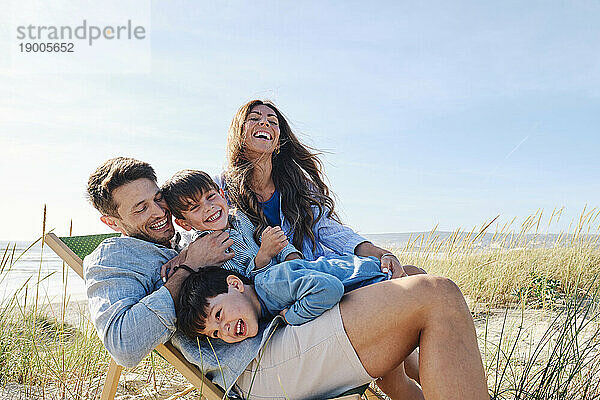 Playful family embracing father sitting on chair at beach