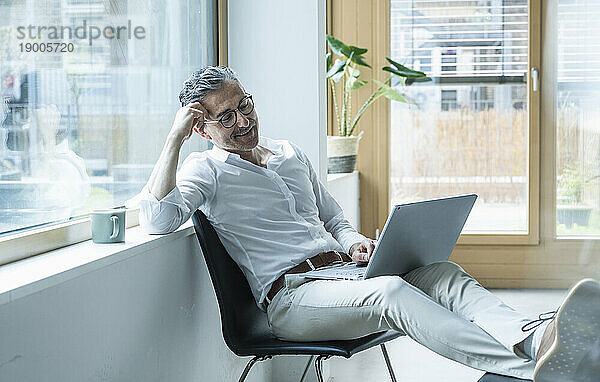 Selbstbewusster Geschäftsmann arbeitet mit Laptop am Fenster im Büro