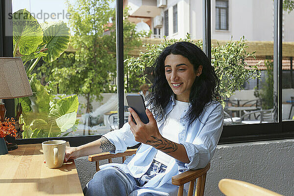 Lächelnde Frau  die auf einem Stuhl im Café sitzt und ihr Smartphone benutzt