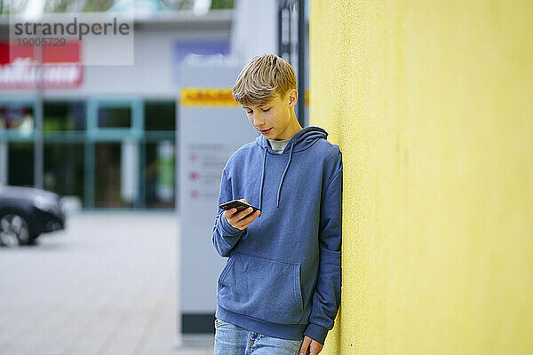 Blonder Junge lehnt an gelber Wand und benutzt Mobiltelefon