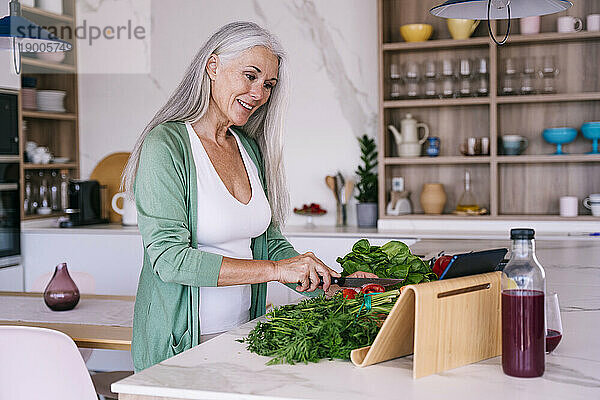 Lächelnde reife Frau schaut sich Tutorial an und bereitet zu Hause Essen zu