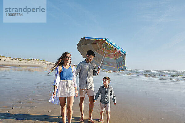 Glückliche Familie  die an einem sonnigen Tag mit Sonnenschirm am Strand spaziert