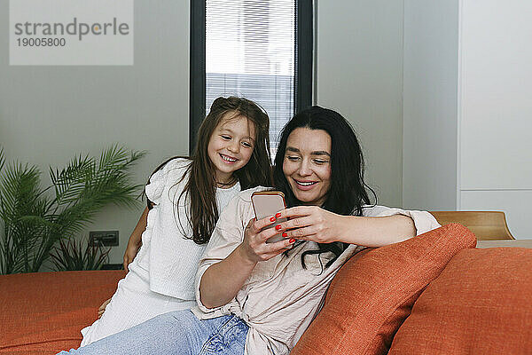 Smiling woman sharing smart phone with daughter at home