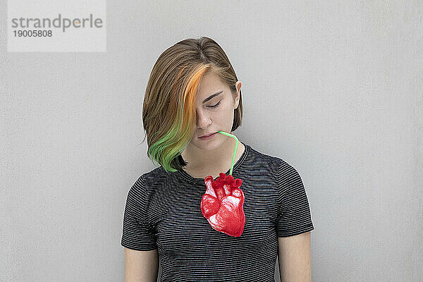 Teenage girl with dyed hair drinking from heart against gray background