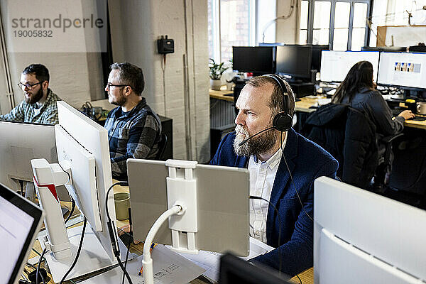 Reifer Geschäftsmann trägt Headset und arbeitet mit Kollegen am Schreibtisch am Computer