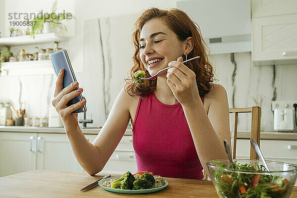 Glückliche junge Frau benutzt Smartphone und isst Salat am Esstisch