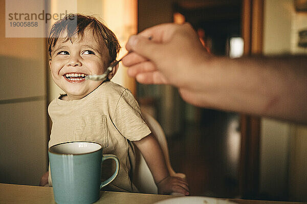 Vater füttert lachenden Sohn zu Hause mit Frühstück