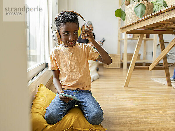 Fröhlicher Junge mit kabellosen Kopfhörern  der zu Hause auf einem Kissen sitzt
