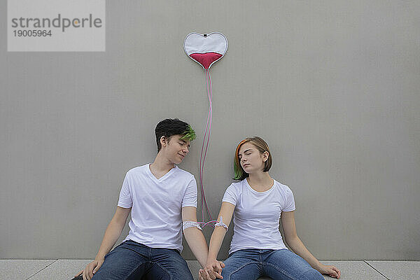 Teenage couple connected with IV drips relaxing in front of gray wall