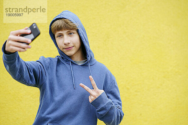Lächelnder Junge zeigt eine Friedenszeichen-Geste und macht ein Selfie mit dem Smartphone