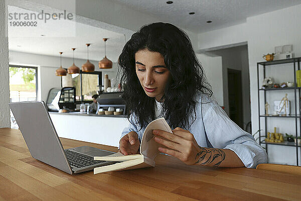 Frau liest Buch mit Laptop auf Tisch im Café