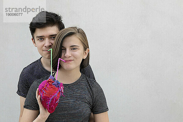 Lächelndes Teenager-Paar trinkt gemeinsam aus Model-Herz vor grauem Hintergrund