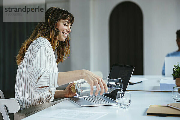 Glückliche Geschäftsfrau gießt Wasser in Glas am Schreibtisch