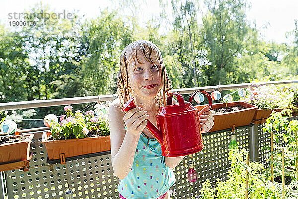 Smiling girl standing and holding red watering can in balcony