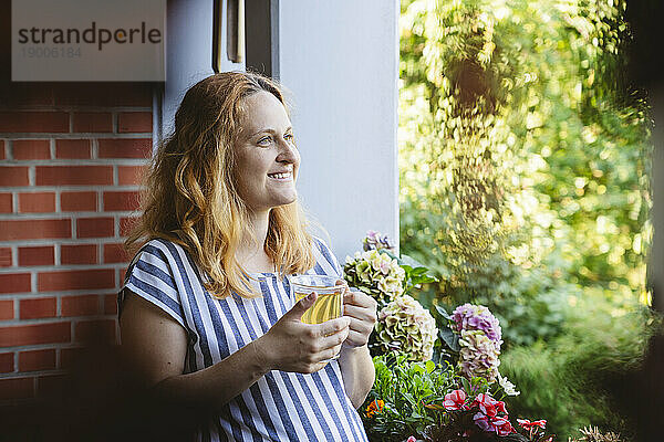 Nachdenkliche schwangere Frau hält eine Tasse Kräutertee auf dem Balkon