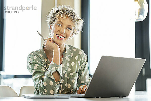 Glückliche blonde Geschäftsfrau sitzt mit Laptop am Tisch