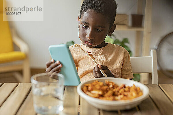 Junge isst Essen und benutzt Smartphone am Esstisch