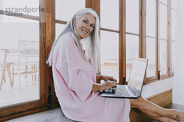 Lächelnde Frau mit Laptop sitzt auf der Fensterbank