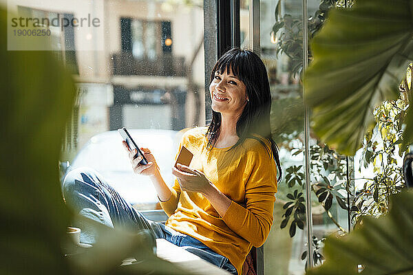 Glückliche Frau mit Kreditkarte und Mobiltelefon sitzt im Café