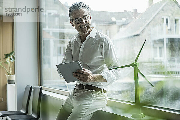 Lächelnder reifer Geschäftsmann mit Tablet-PC  der im Büro das Modell einer Windkraftanlage betrachtet