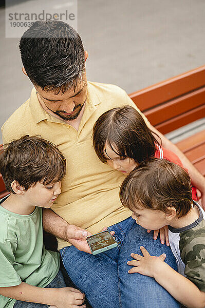 Vater teilt Smartphone mit Söhnen  die auf Bank im Park sitzen