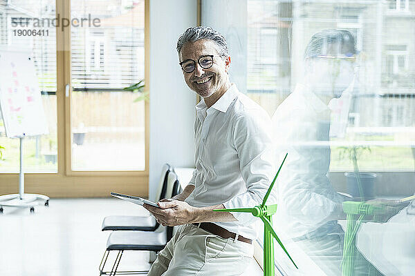 Glücklicher reifer Geschäftsmann mit Tablet-PC und Windturbinenmodell im Büro