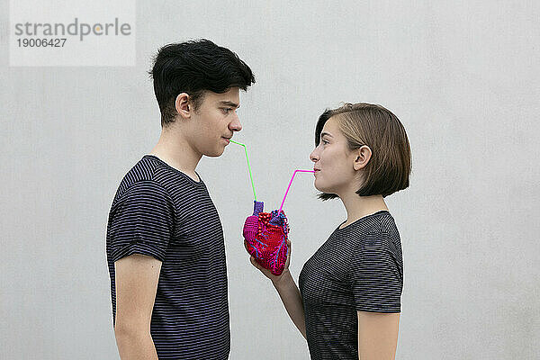 Teenage couple drinking together from model heart against gray background