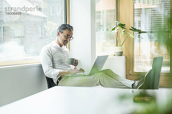 Selbstbewusster Geschäftsmann mit Kaffeetasse und Laptop bei der Arbeit im Büro