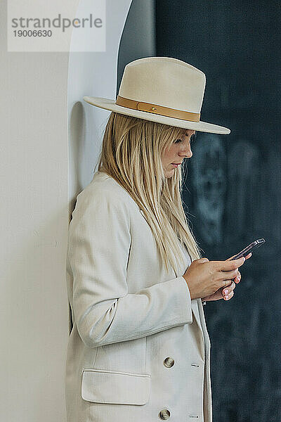 Blonde Frau benutzt Smartphone und steht an der Wand