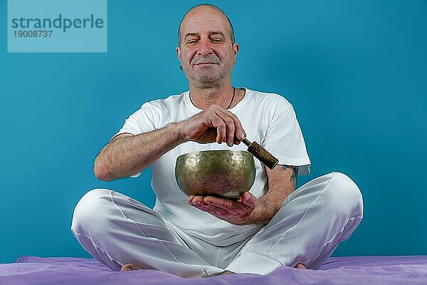 Weiß gekleideter Mann  der im Zustand der Meditation eine tibetische Klangschale anschlägt