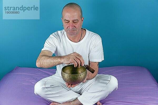 Weiß gekleideter Mann  der im Zustand der Meditation eine tibetische Klangschale anschlägt