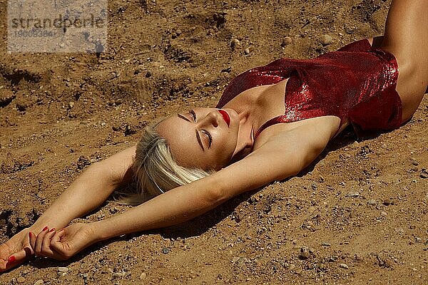 Schöne blonde Frau in einem roten sexy Kleid auf dem Hintergrund des Himmels und Sand. Schönheit des Körpers und des Gesichts