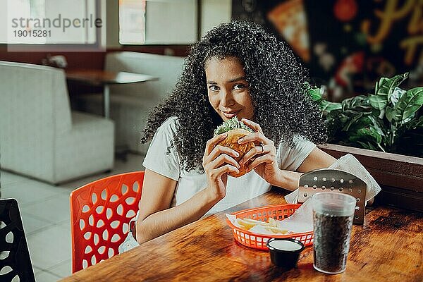 Porträt eines Afro Mädchens  das einen Hamburger in einem Restaurant hält. Schönes lateinisches Mädchen genießt einen leckeren Hamburger in einem Restaurant