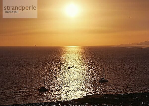 Wunderschöne Meereslandschaft bei Sonnenuntergang. Niedrige Sonne in dunstigen Himmel und Sonne Blendung auf ruhigem Meer Wasser. Silhouette von kleinen Segelbooten gehen  um Meer  Sonnenlicht Streifen  dunkle Küste im Vordergrund. Sommer heiße Tage  Urlaub  Freizeit  romantisches Segeln