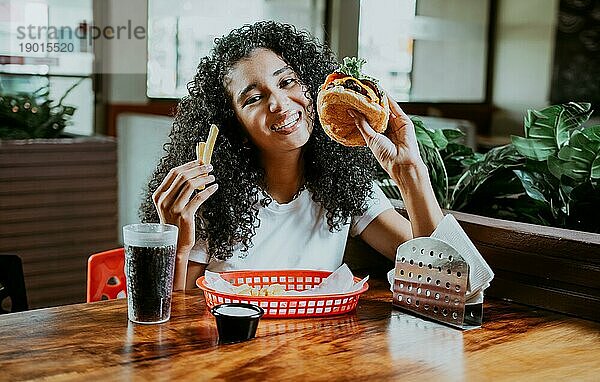 Afro junge Frau genießt einen Hamburger mit Pommes in einem Restaurant. Glückliches Mädchen genießt einen Hamburger in einem Restaurant