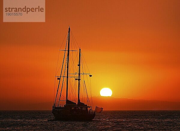 Silhouette eines Segelboots mit heruntergelassenen Segeln gegen die Sonne bei Sonnenuntergang  Sonnenblendung auf dem Meer. Romantische Meereslandschaft. Segelboot fährt ins Meer  Rückansicht  Sonnenuntergang im dunstigen Hintergrund. Meditatives Bild