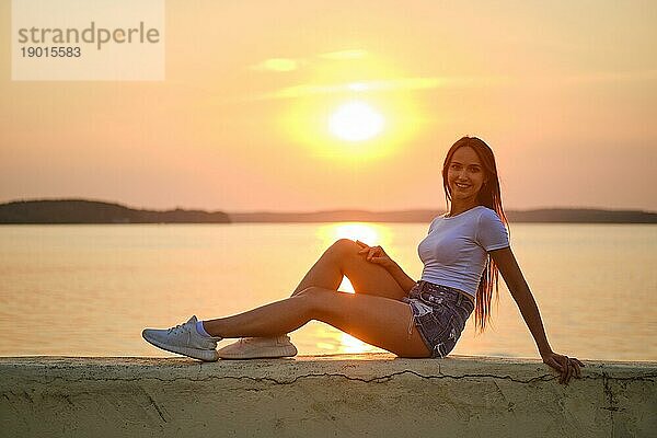 Lächelnde Frau sitzt bei Sonnenuntergang auf einem Steg