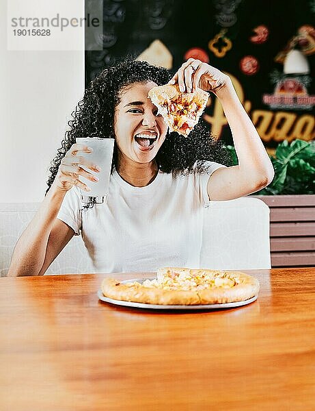 Glückliche afrohaarige Frau zeigt ein Stück Pizza in einem Restaurant. Lächelnde Afro Frau  die eine Pizza in einem Restaurant genießt