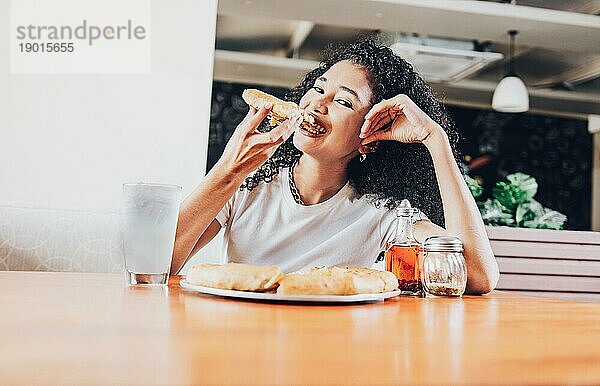 Lächelnde afro haarige Frau  die eine Pizza in einem Restaurant genießt. Glückliche afrohaarige Frau isst Pizza in einem Restaurant