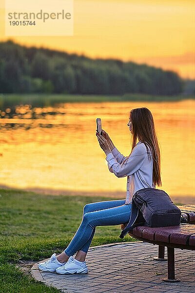 Junge Frau fotografiert sitzend am Ufer eines Flusses bei Sonnenuntergang