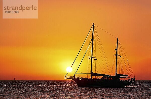 Silhouette von Segelboot oder Schoner mit Segeln nach unten gegen die Sonne bei Sonnenuntergang  Sonne Blendung auf Meerwasser. Romantische Meereslandschaft  Sonne berühren Vorsegel Taue. Klarer orangefarbener Himmel. Meditatives Bild von Sonnenuntergang und Boot